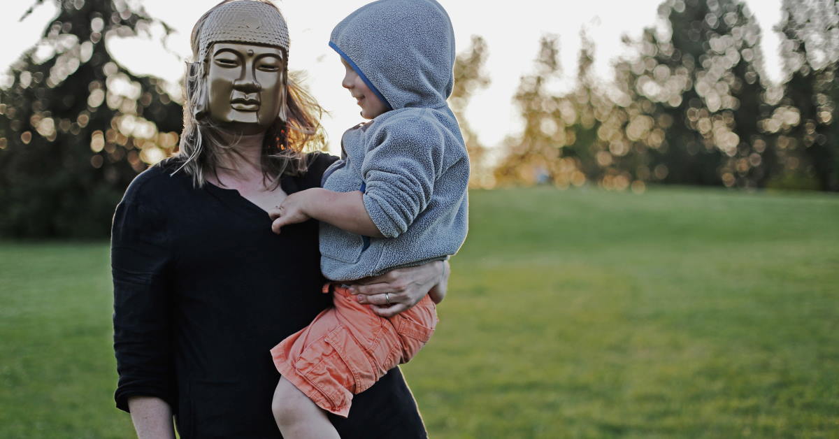 parent with mask carrying toddler with hoodie on grass field