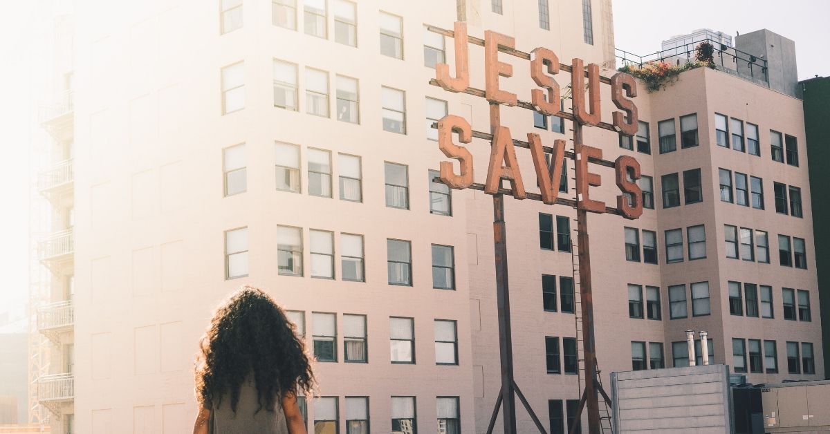 photo of a woman sitting on a wall overlooking skyrises beside a sign which says "jesus saves"