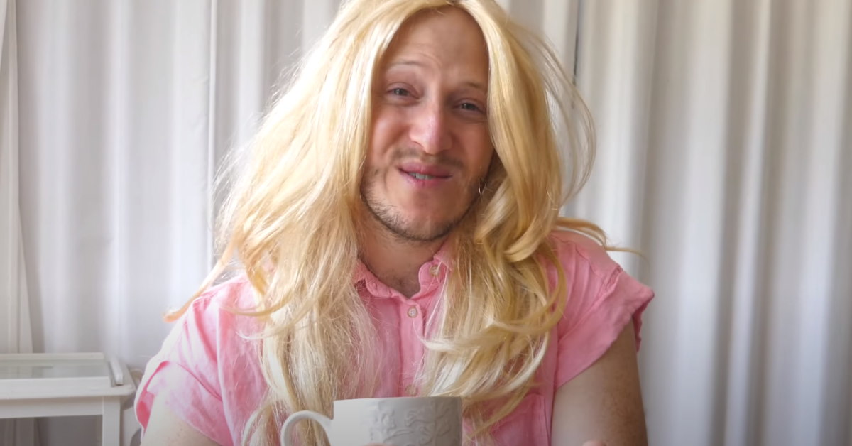Bearded comedian dressed as female in pink blouse and blonde wig holding cup of tea