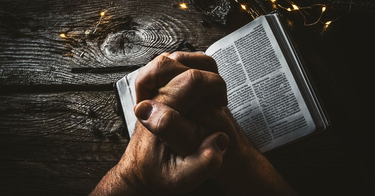 hands clasped in prayer over a bible on a dark timber table
