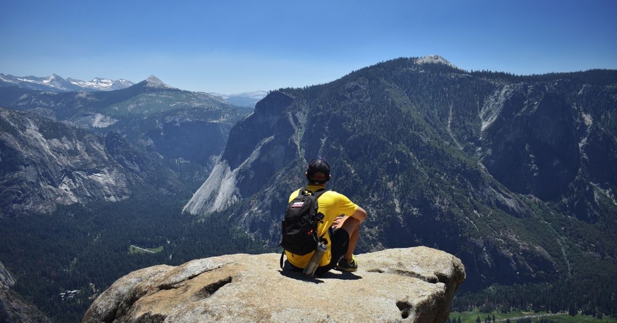 man in a yellow shirt sitting on a rock ledge ovelooking a mountain range