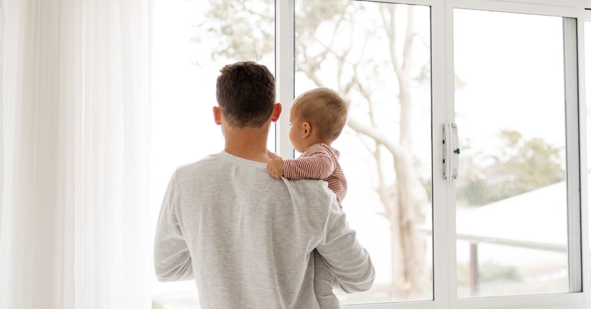 young dad holding baby standing at a window inside the house