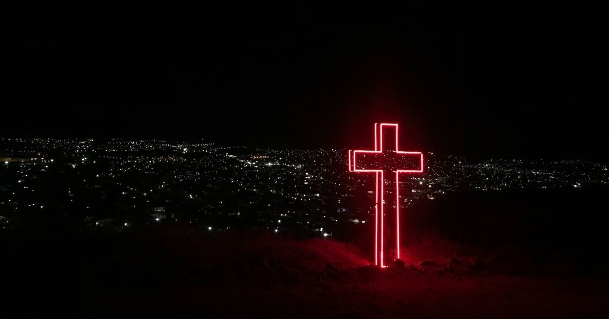 neon pink cross lit up in the foreground. in the background are city lights at night time