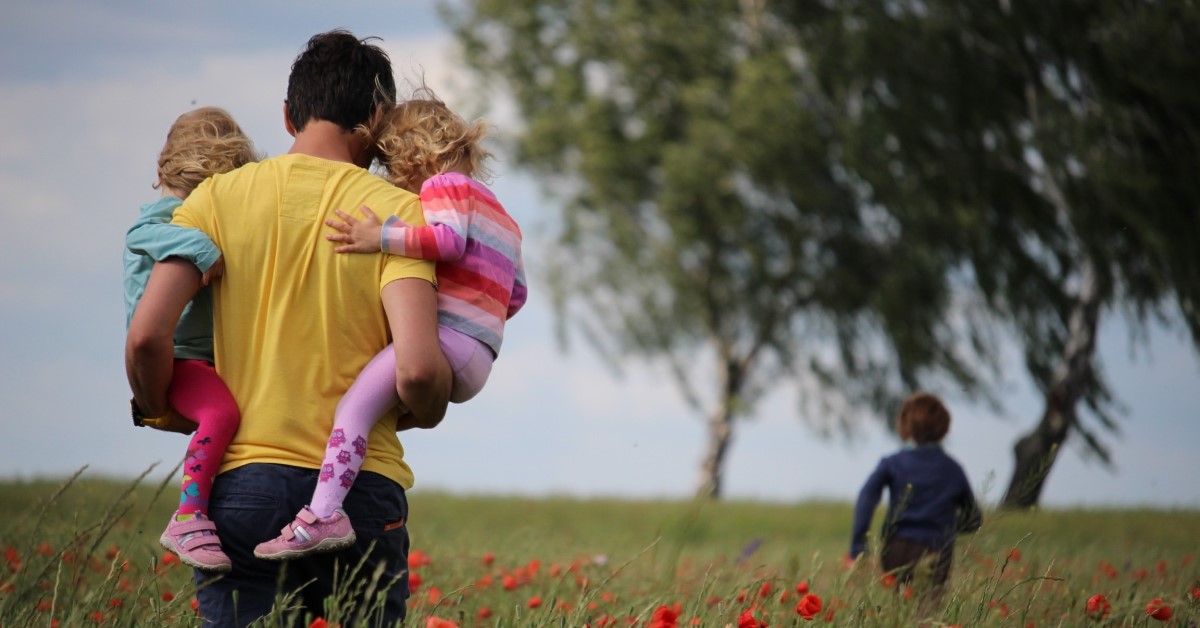 father carrying a child in each arm through a field with another child running through field ahead